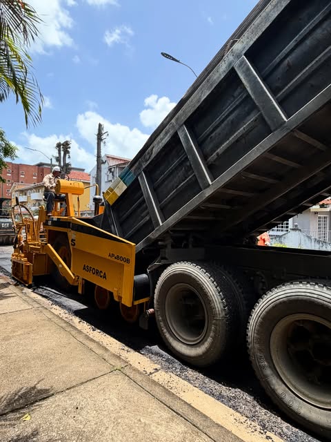Camión volqueta descargando mezcla en una pavimentadora sobre una calle urbana